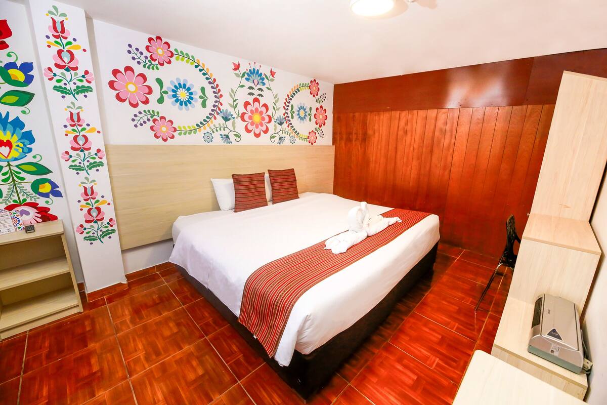 Une chambre d’hôtel avec un plancher en bois, un lit recouvert de draps blancs et d’une couverture à rayures rouges, une tête de lit en bois et un mur décoré de motifs floraux colorés.