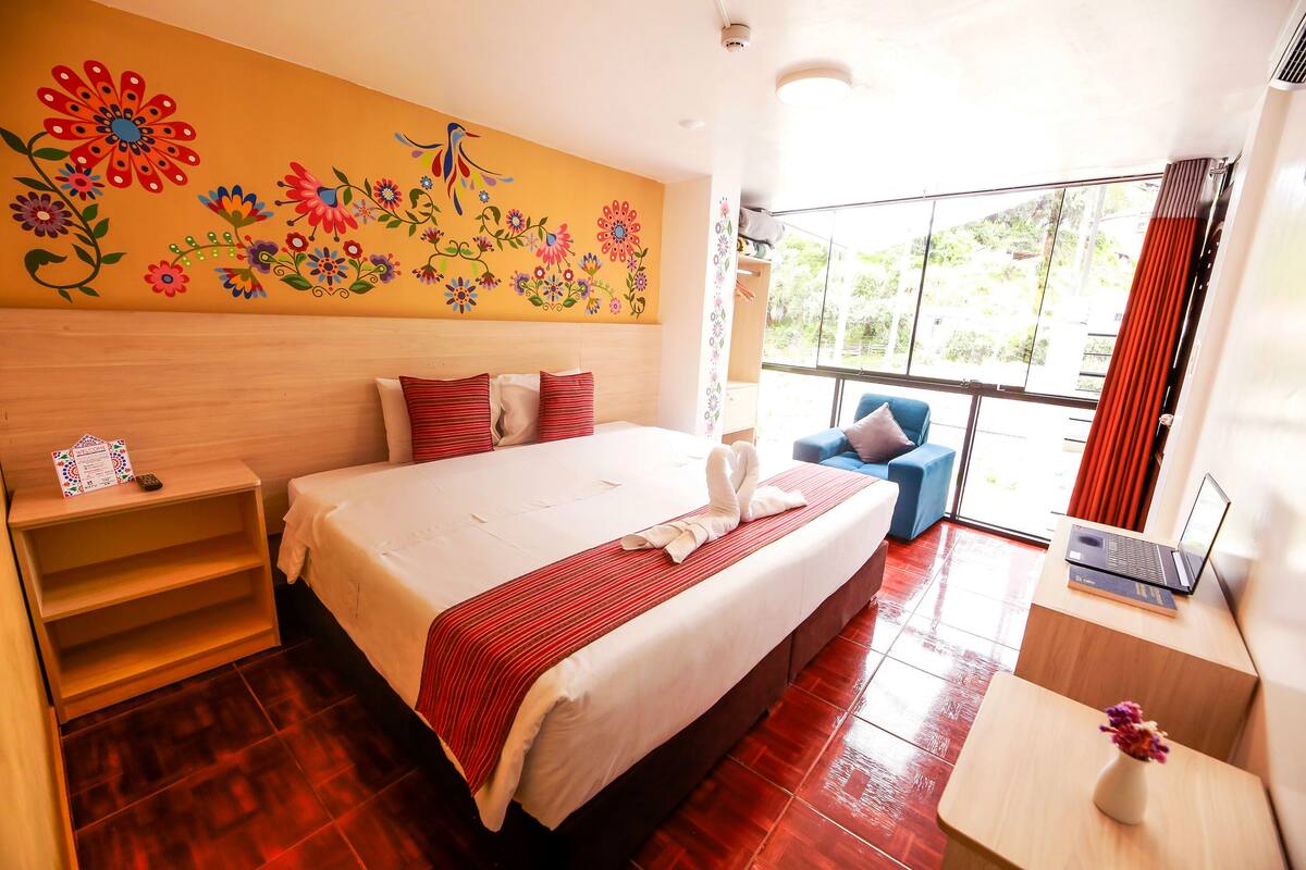 Une chambre d’hôtel comprenant un lit, un bureau avec un ordinateur portable, une chaise, une fenêtre avec des rideaux et un mur décoré d’œuvres d’art florales.
