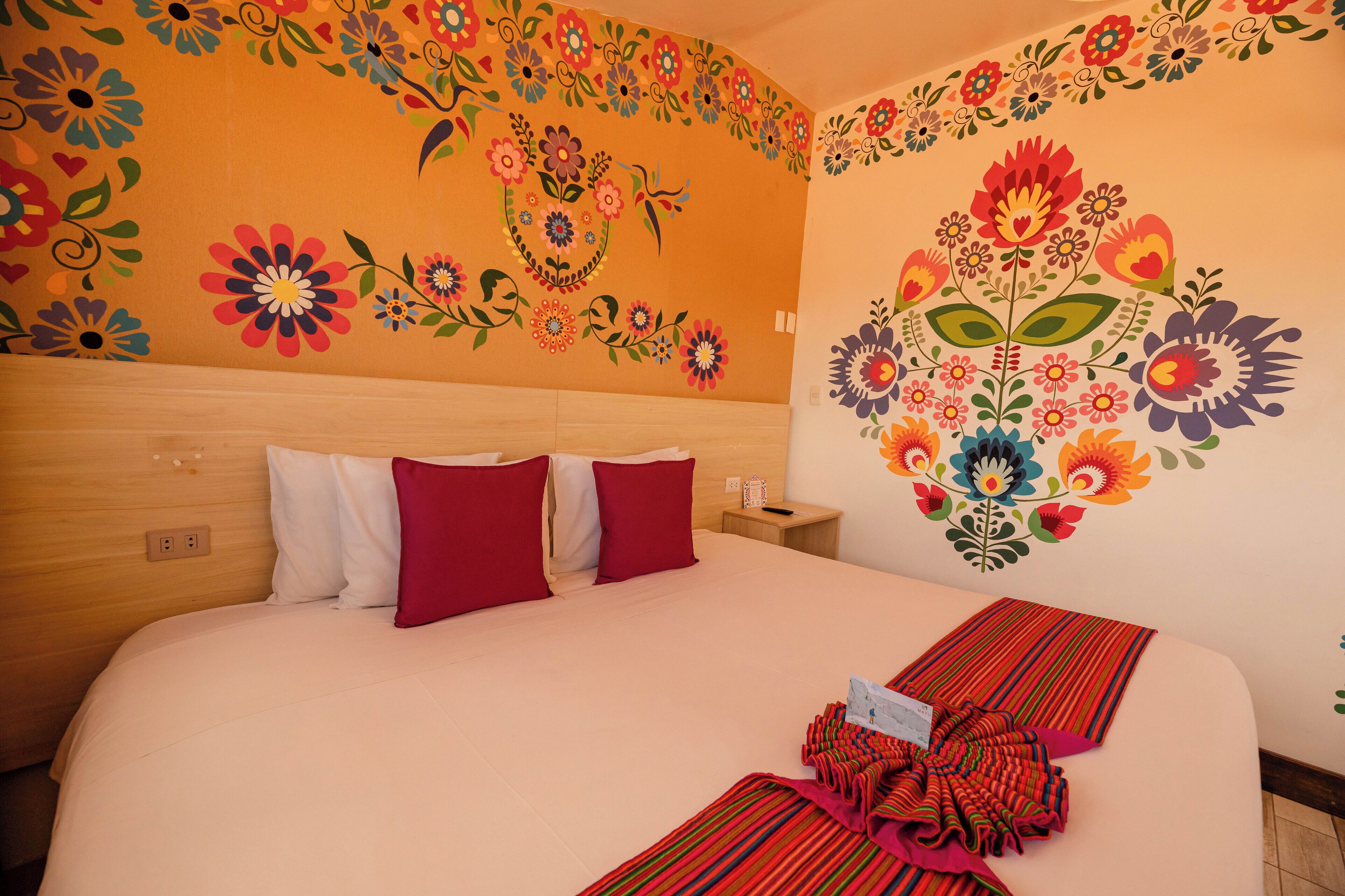 Habitación de hotel con una cama grande, almohadas rojas y blancas, cabecera de madera y un mural floral en la pared.