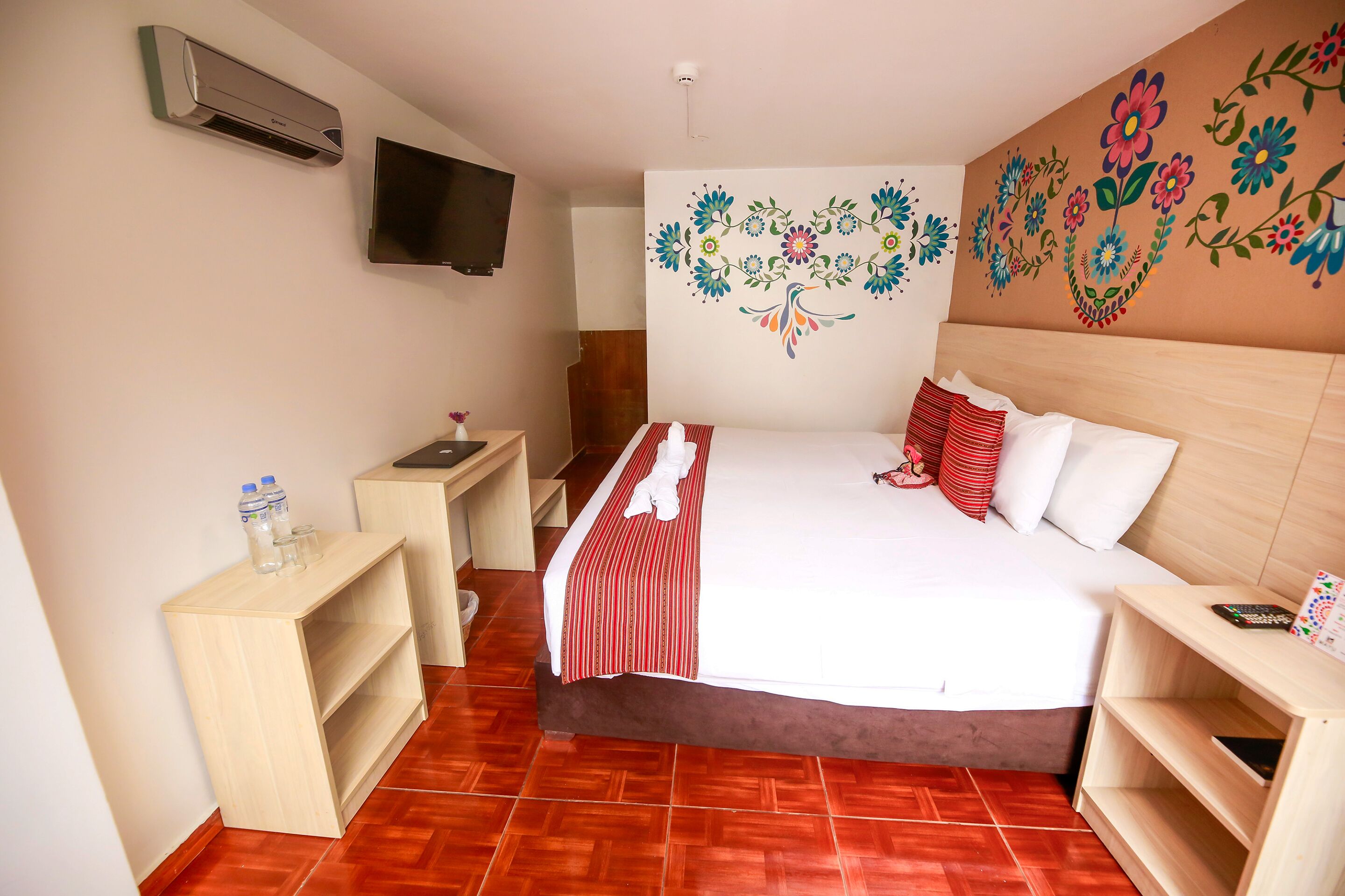 A hotel room with a bed, a TV mounted on the wall, a wooden nightstand, and a wall decoration.