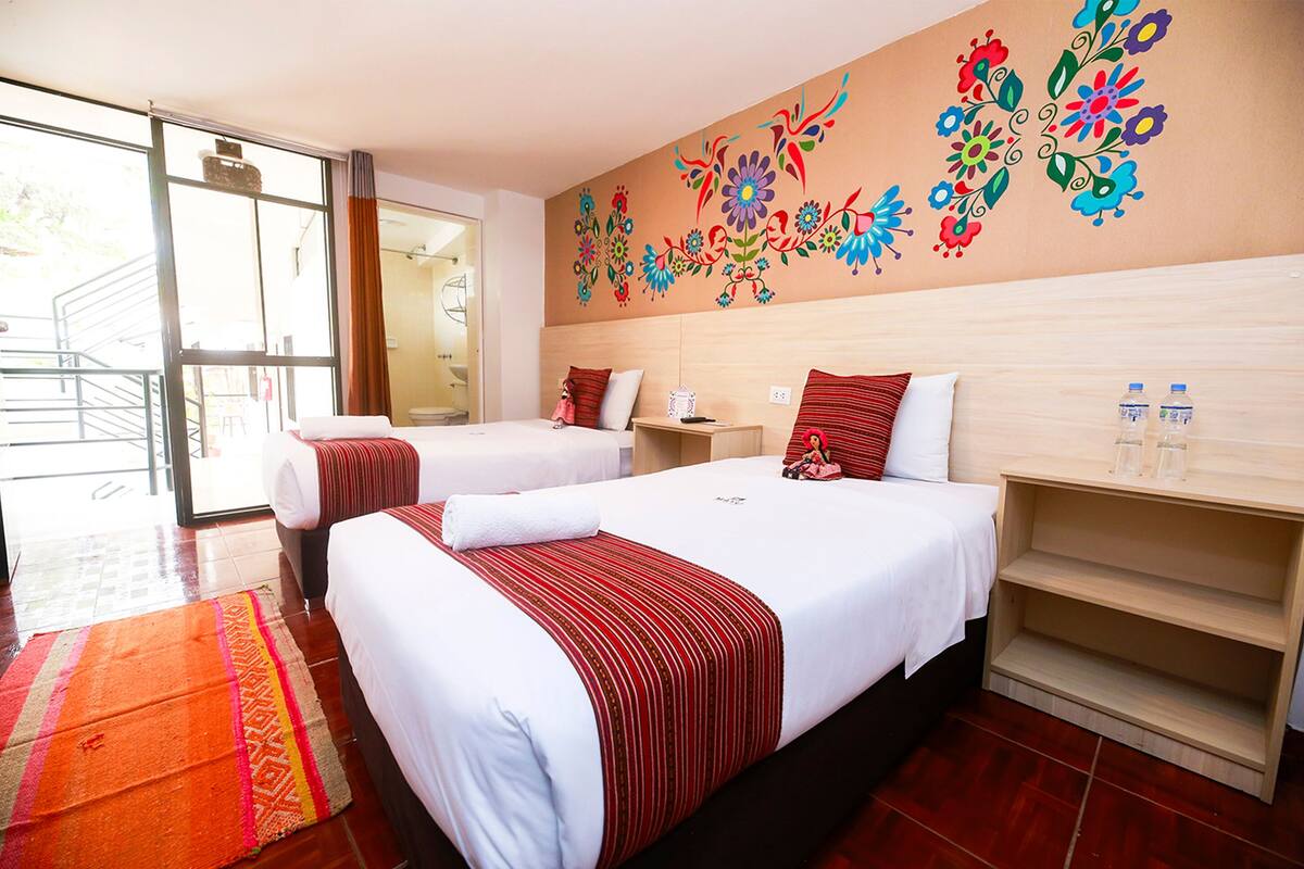 Une chambre d’hôtel avec deux lits, une grande fenêtre, une tête de lit en bois et une fresque murale aux motifs floraux et aux couleurs vives.