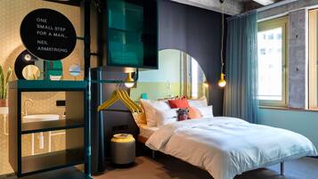 A modern hotel room with a bed, bedside table, mirror, and a view of the city.