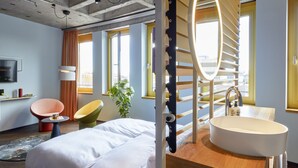 Una habitación de hotel moderna con una cama grande, un espejo redondo, un tocador de madera con un lavabo, una silla roja y una planta en maceta.