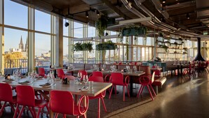 Un restaurante espacioso con techos altos, conductos expuestos y grandes ventanales que ofrecen vistas a la ciudad. Las mesas están preparadas con sillas rojas y platos vacíos, señal de que esperan a los comensales.