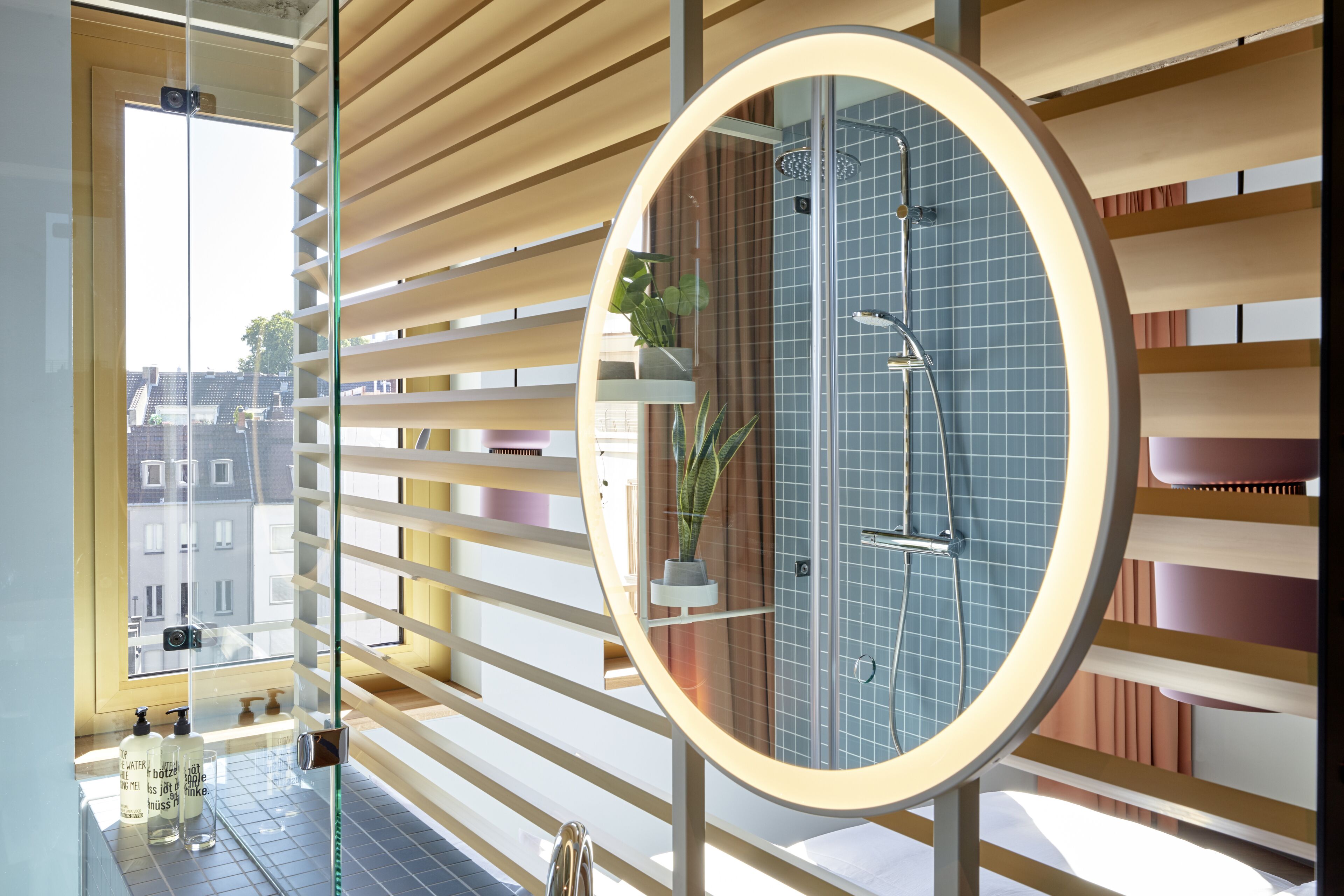 A modern bathroom with a glass shower enclosure, a circular mirror, and a view of the city through large windows.