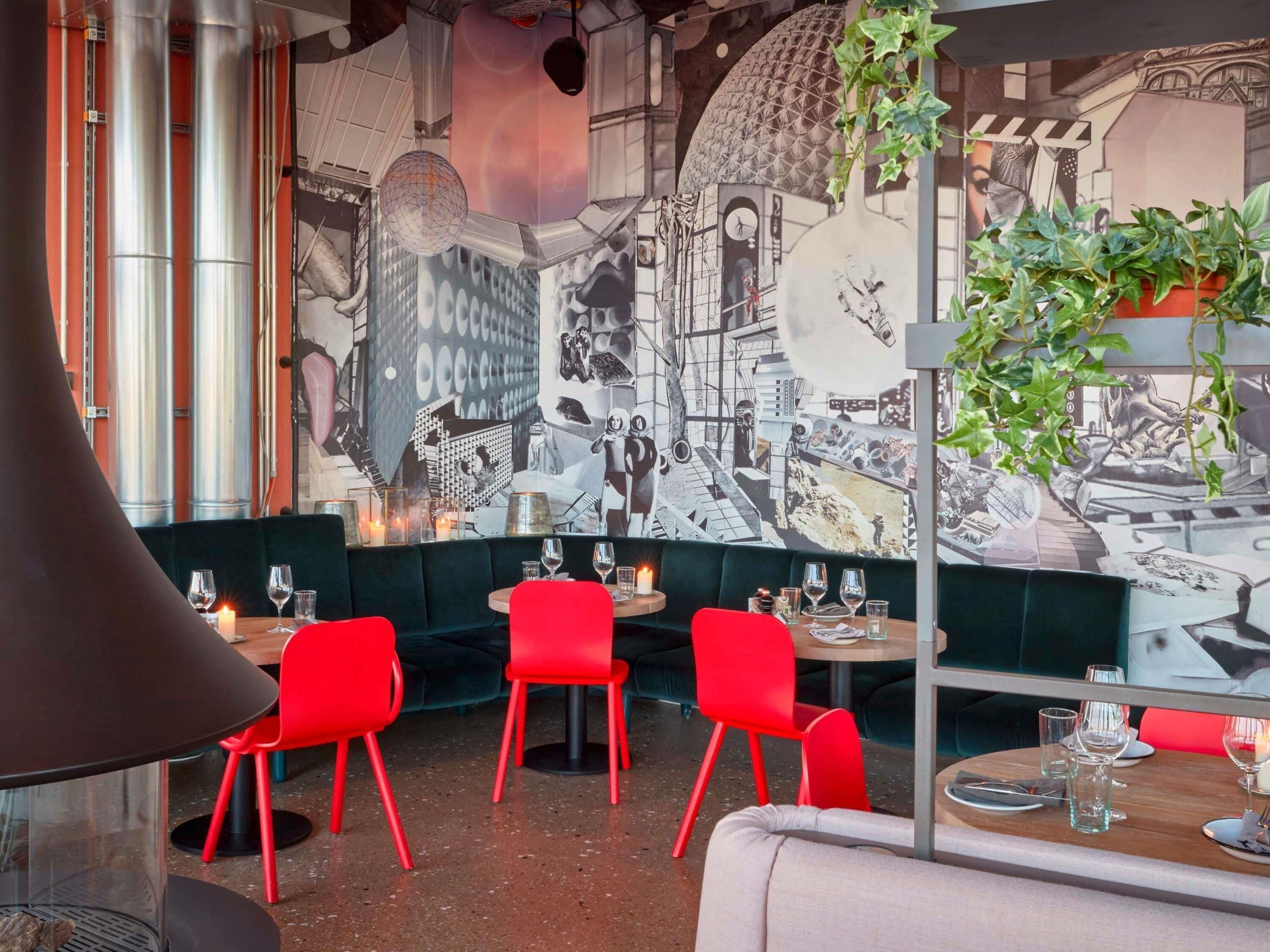 Interior de restaurante con sillas rojas, un mural de un paisaje urbano y una planta colgante.