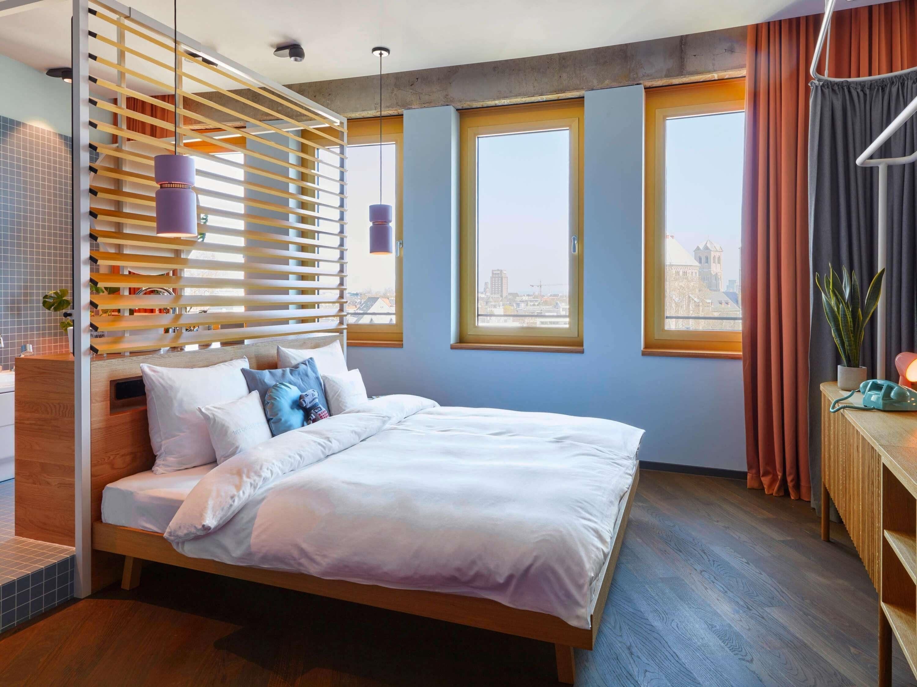 Ein modernes Hotelzimmer mit einem großen Bett, einem hölzernen Kopfteil, Blick auf die Stadt durch die Fenster und einem kleinen Schreibtisch mit Telefon.
