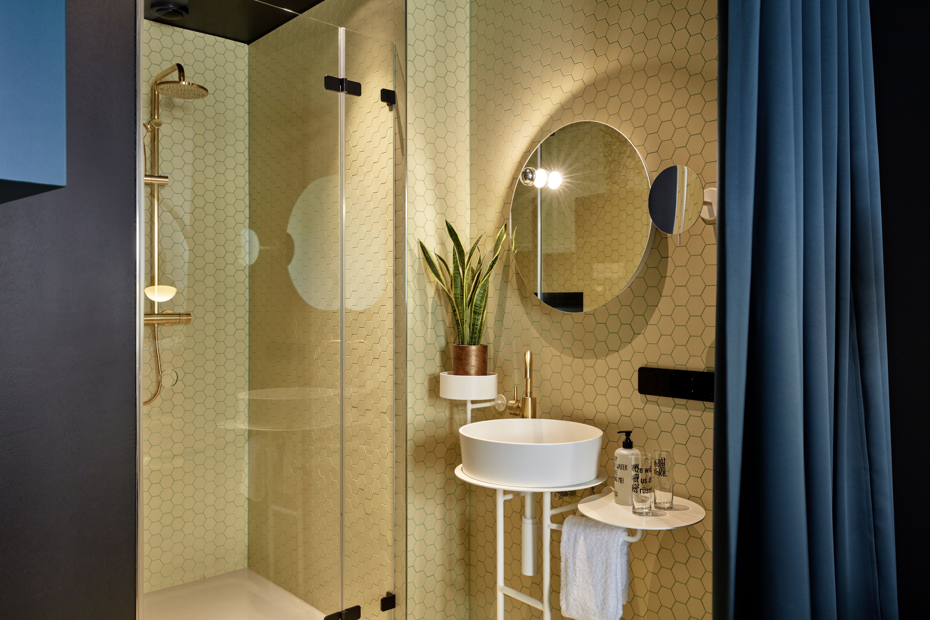 A modern bathroom with a glass-enclosed shower, a round mirror, a white sink, and a small shelf with toiletries.