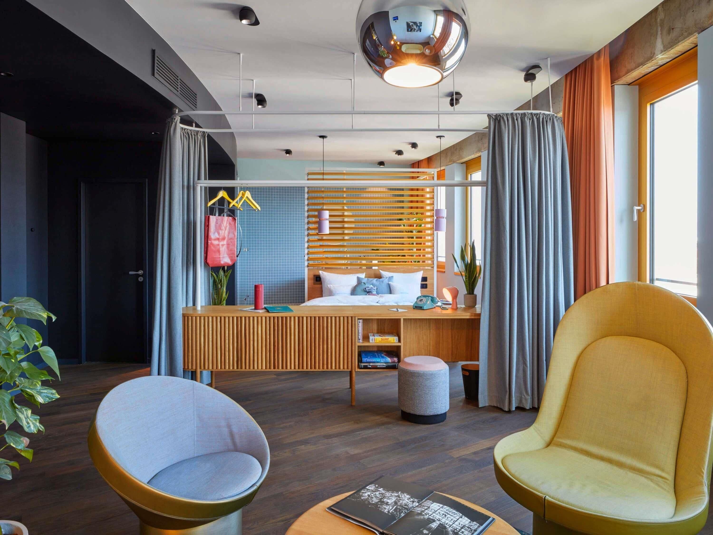 Habitación de hotel moderna con cama, mesita de noche, dos sillas, un espejo y una planta en maceta.