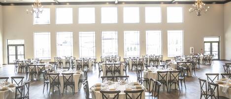 A spacious dining hall with multiple tables set for a meal, large windows, and chandeliers.