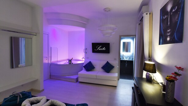 A modern hotel room with a large bathtub, a sofa with cushions, a flat-screen TV, and a vase with flowers on a console table. - Maschio Angioino Suite (Naples)