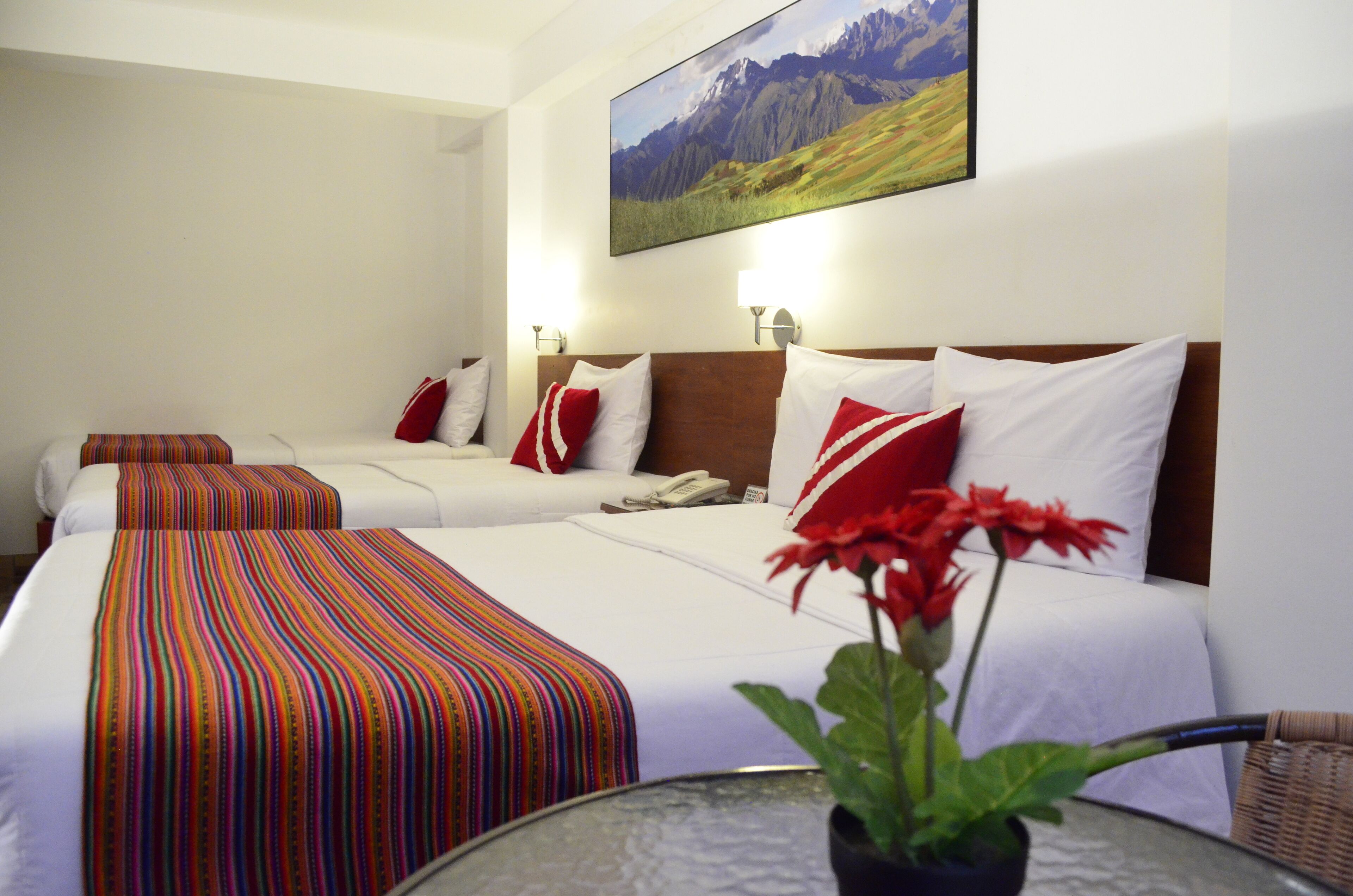 Habitación de hotel con dos camas, cada una con una colcha a rayas y almohadas rojas. En la pared hay un cuadro de un paisaje montañoso, y sobre la mesa, un jarrón con flores rojas.