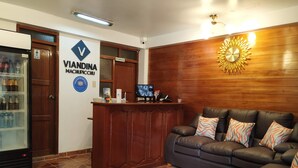 A reception area with a wooden reception desk, a couch with patterned cushions, a refrigerator stocked with beverages, and a clock on the wall. - Viandina Machupicchu (Machu Picchu)