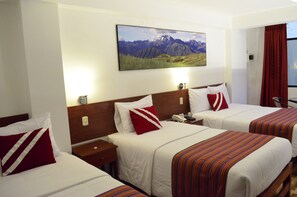 Habitación de hotel con dos camas, cada una con una colcha blanca y almohadas rojas y blancas. Tiene un cabecero de madera, una mesita de noche con una lámpara y un cuadro colgado en la pared que muestra un paisaje de montaña.