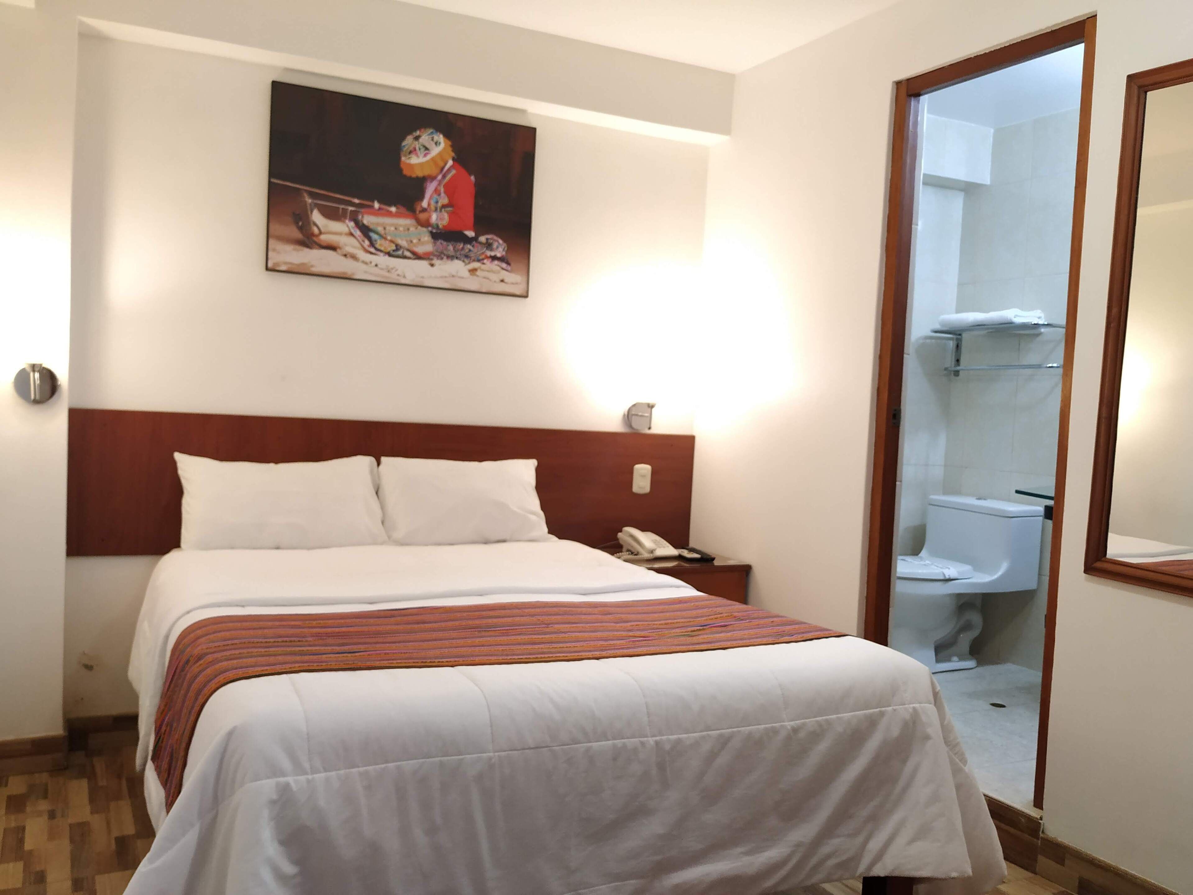 A hotel room with a bed, a nightstand with a phone, a painting on the wall, and a bathroom visible through an open door.