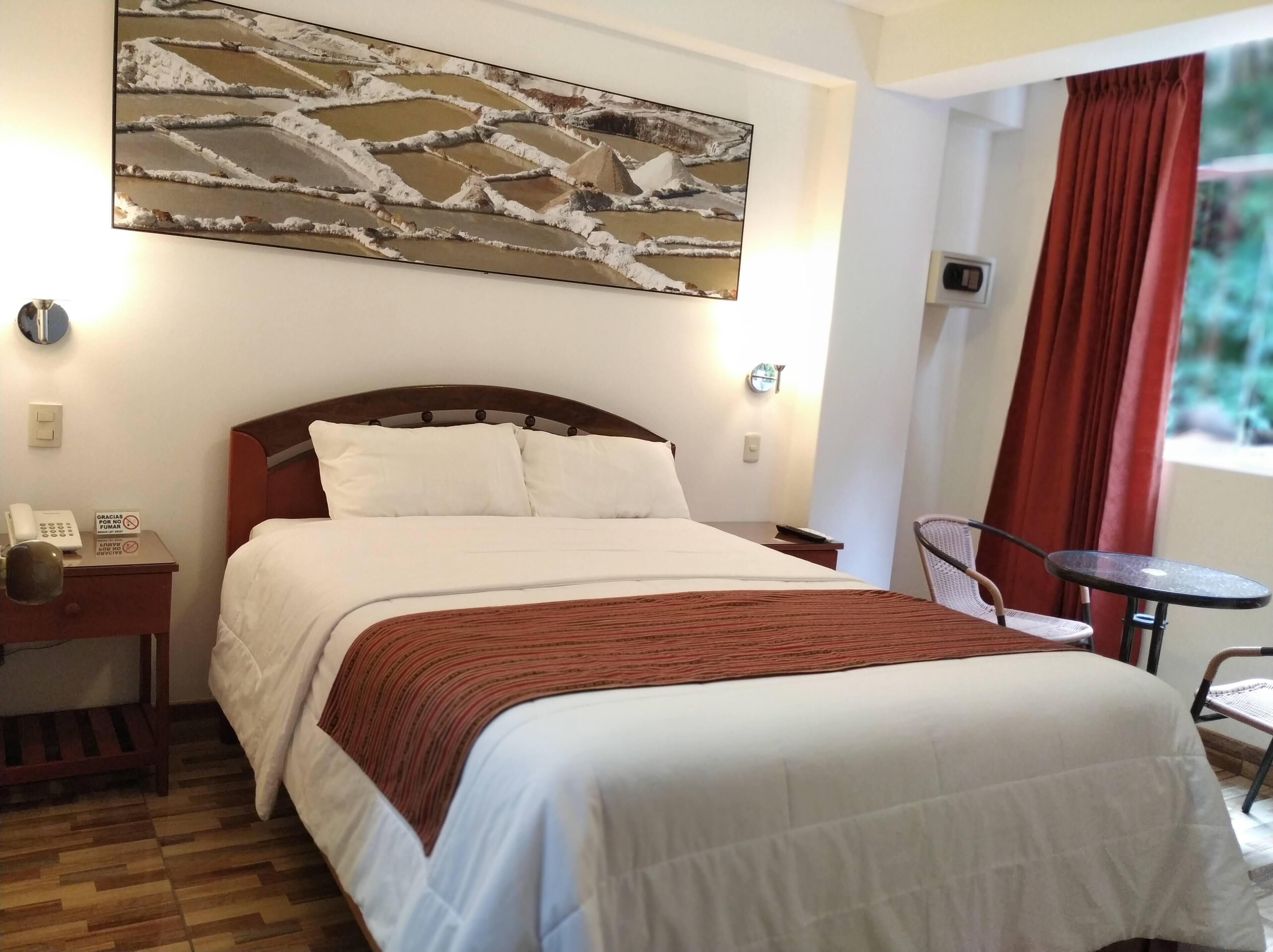 A hotel room with a bed, a bedside table with a phone, a chair, a small table, and a window with curtains.