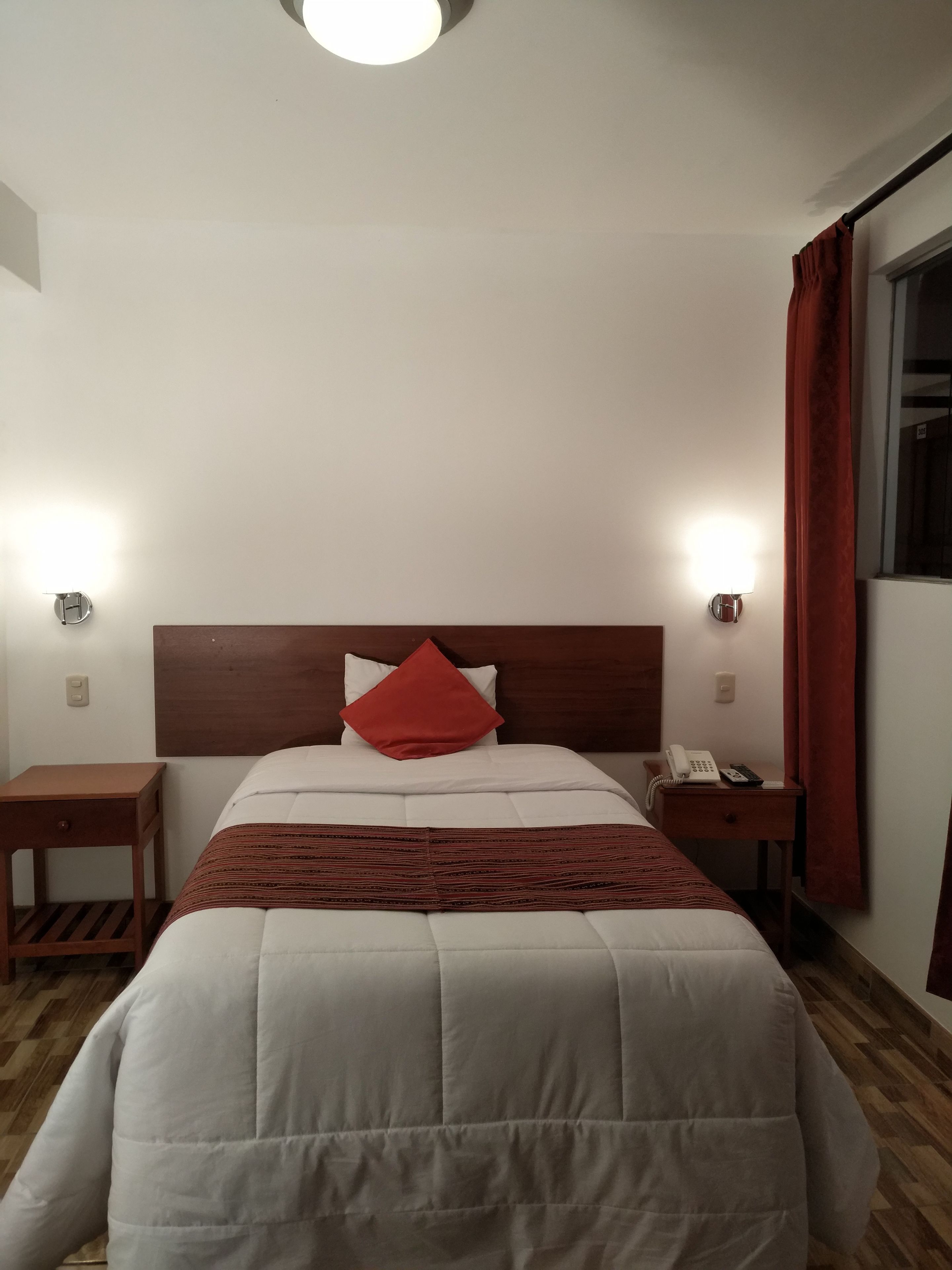 A hotel room with a bed, two bedside tables, a nightstand, a red curtain, and a window.