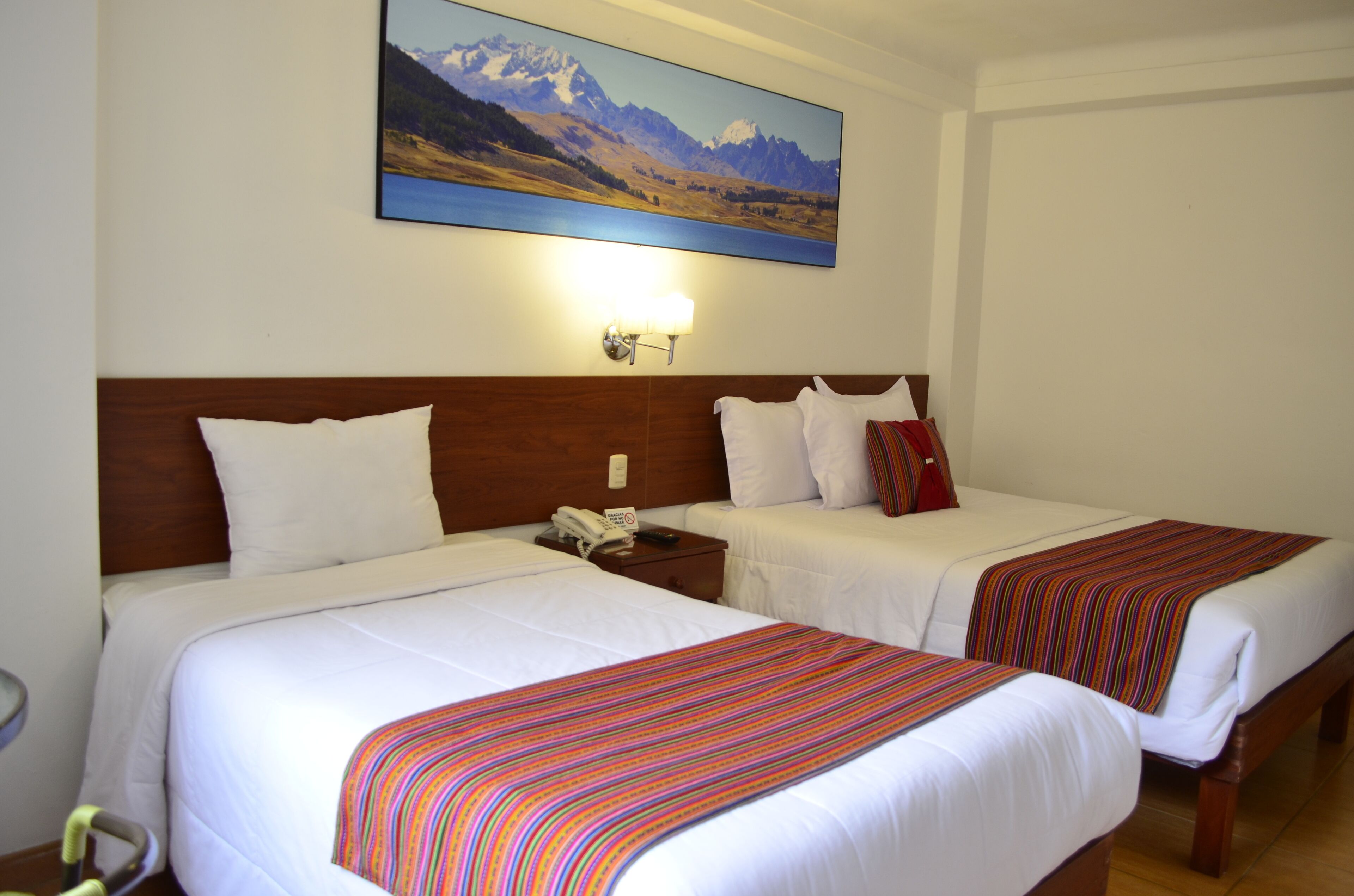 Una camera d'albergo con due letti, una testiera in legno, biancheria da letto bianca, una coperta a righe, un quadro raffigurante montagne e un comodino con telefono e telecomando.
