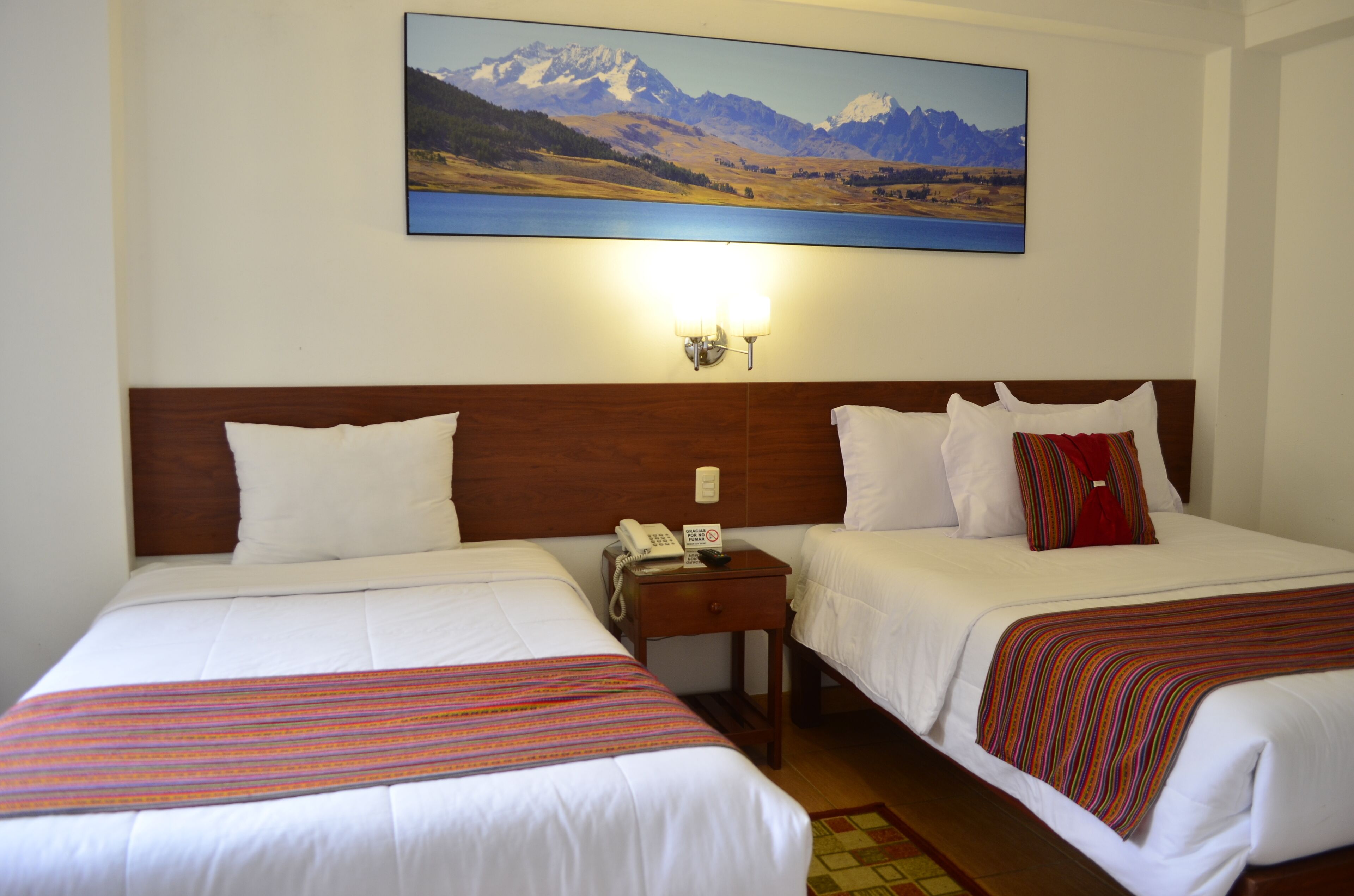 A hotel room with two beds, a wooden headboard, a painting of a mountain landscape, and a nightstand with a phone and a sign.