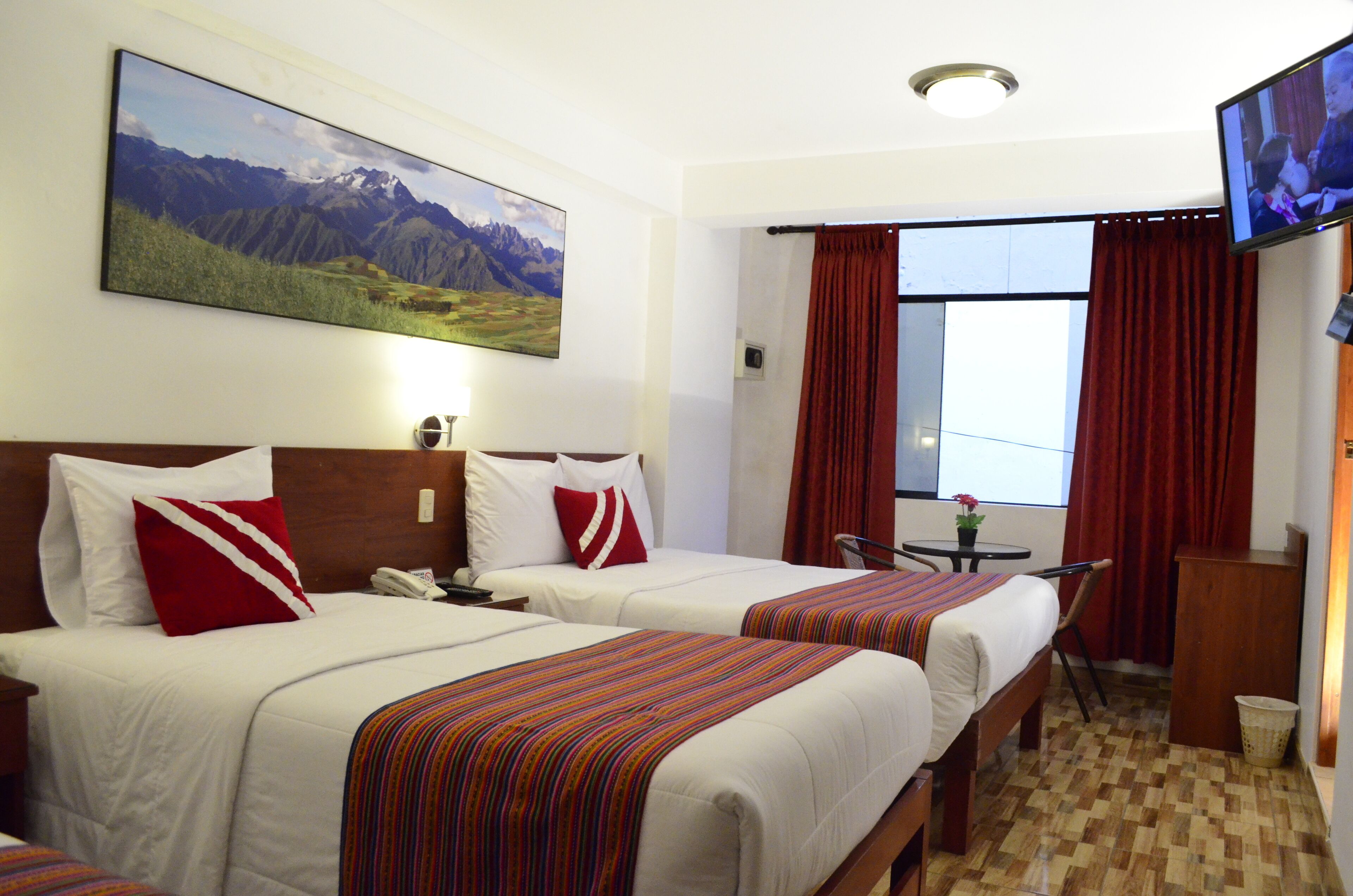 A hotel room with two beds, a TV, a window with red curtains, and a painting of mountains on the wall.
