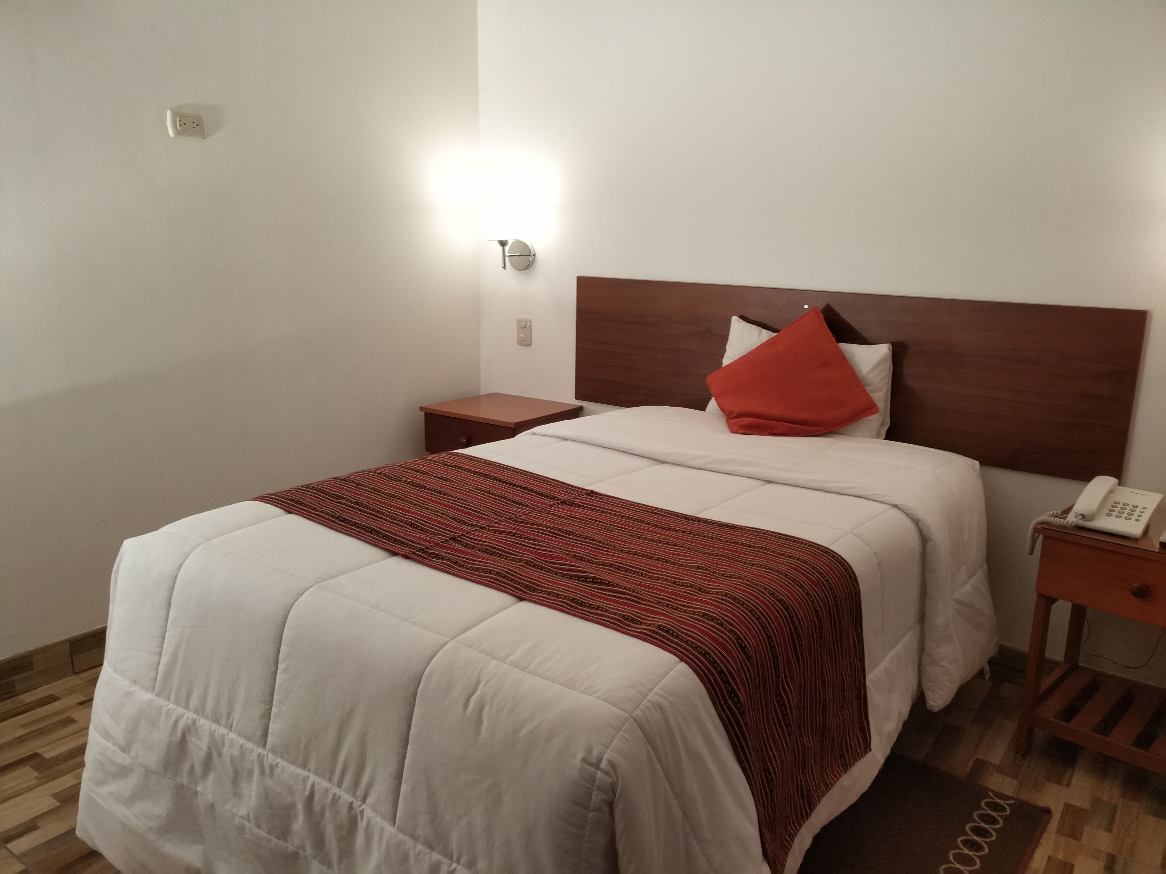 A hotel room with a bed, a nightstand, a telephone, and a wall socket.