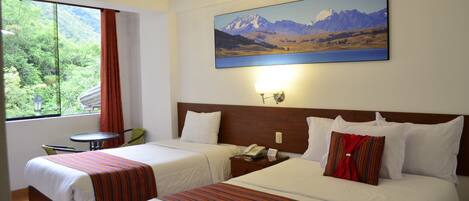 A hotel room with two beds, a window with a view of greenery, a painting of mountains, and a small table with a chair.
