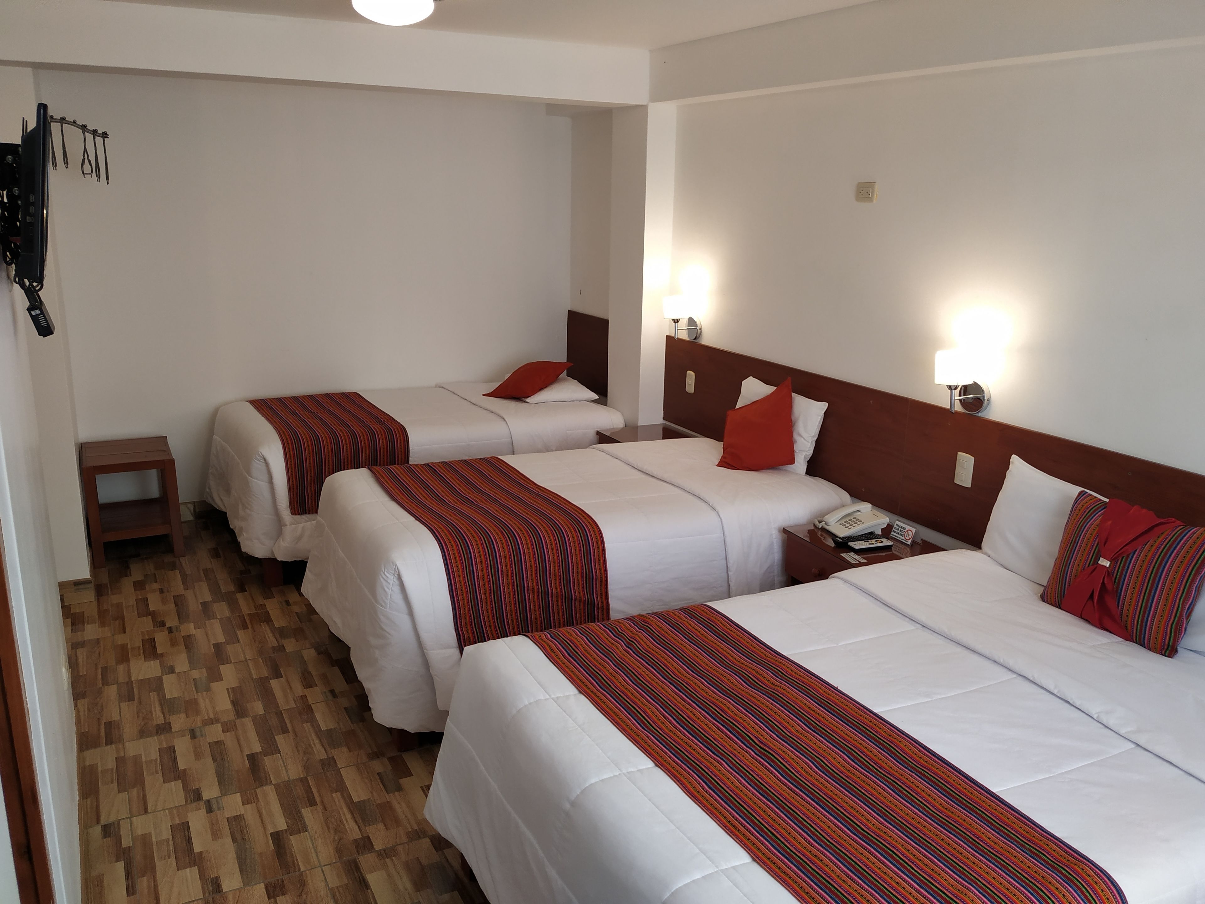 Habitación de hotel con tres camas, cada una con ropa de cama blanca y detalles en rojo. Hay mesitas de noche con lámparas, suelo de madera y un estante pequeño.