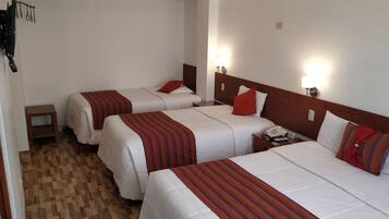 Habitación de hotel con tres camas, cada una con ropa de cama blanca y detalles en rojo. Hay mesitas de noche con lámparas, suelo de madera y un estante pequeño.