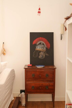 Um quarto com mesa de cabeceira de madeira, quadro com capacete, sonho apanhador vermelho e cama branca.