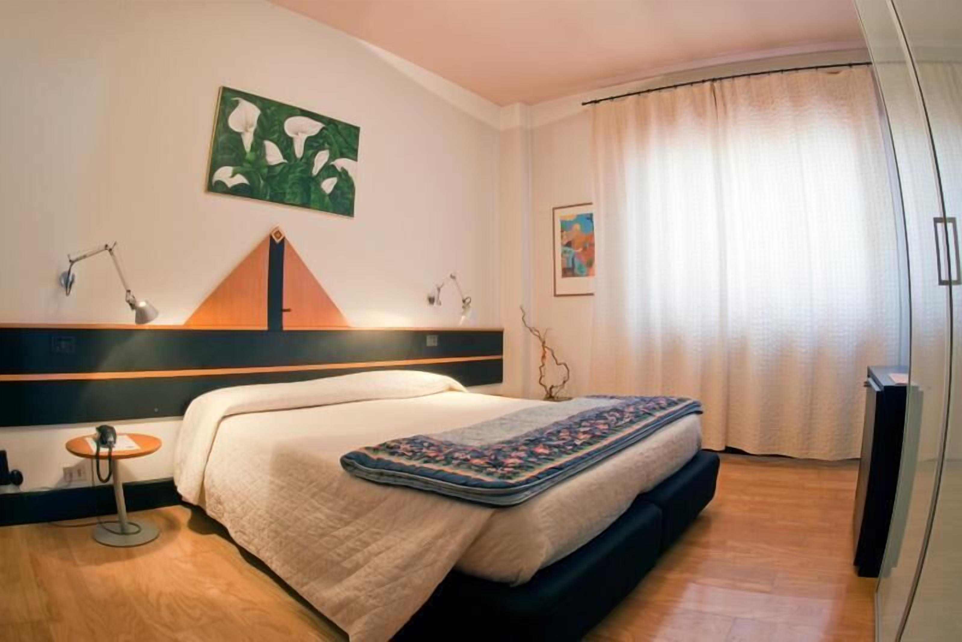 Una camera d'albergo con un letto, una scrivania, una sedia, una lampada e un quadro appeso al muro.