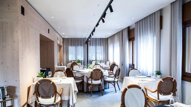 A modern dining area with round tables and wicker chairs, large windows with curtains, and a wooden wall. - Locanda San Lorenzo (Alpago)