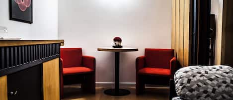 A small, modern room with a wooden floor, a round table with a vase of red flowers, two red chairs, a sofa with a patterned cover, and a black-framed mirror on the wall.