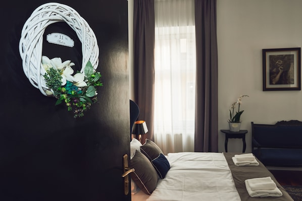 A hotel room with a bed, a dark wall, a white wicker wreath, a framed picture, and a window with curtains. - Shalom Kazimierz (Kraków)