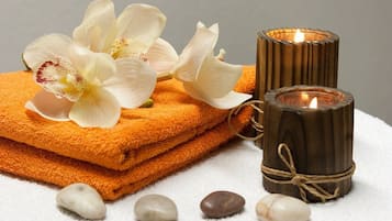 A stack of orange towels, a white flower, a lit candle, and smooth stones on a white surface.