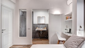 A modern bedroom with a large bed, a bedside desk with a laptop, a shower, and a vanity with a mirror. - Sorrento Lux Suites - Blue (Sorrento)