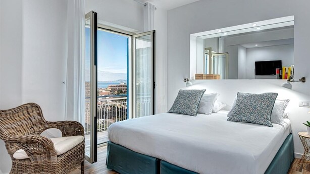 A modern bedroom with a large bed, a wicker chair, a balcony with a city view, and a TV on the wall. - Sorrento Lux Suites - Blue (Sorrento)