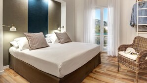 A bedroom with a large bed, two bedside lamps, a wicker chair, and a balcony with curtains. - Sorrento Lux Suites - Blue (Sorrento)