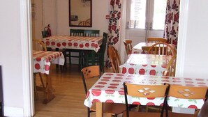 Ein Essbereich mit Tischen, die mit Erdbeer-Muster-Tischdecken bedeckt sind, Holzstühlen und einem braunen Ledersofa mit einem floralen Kissen.