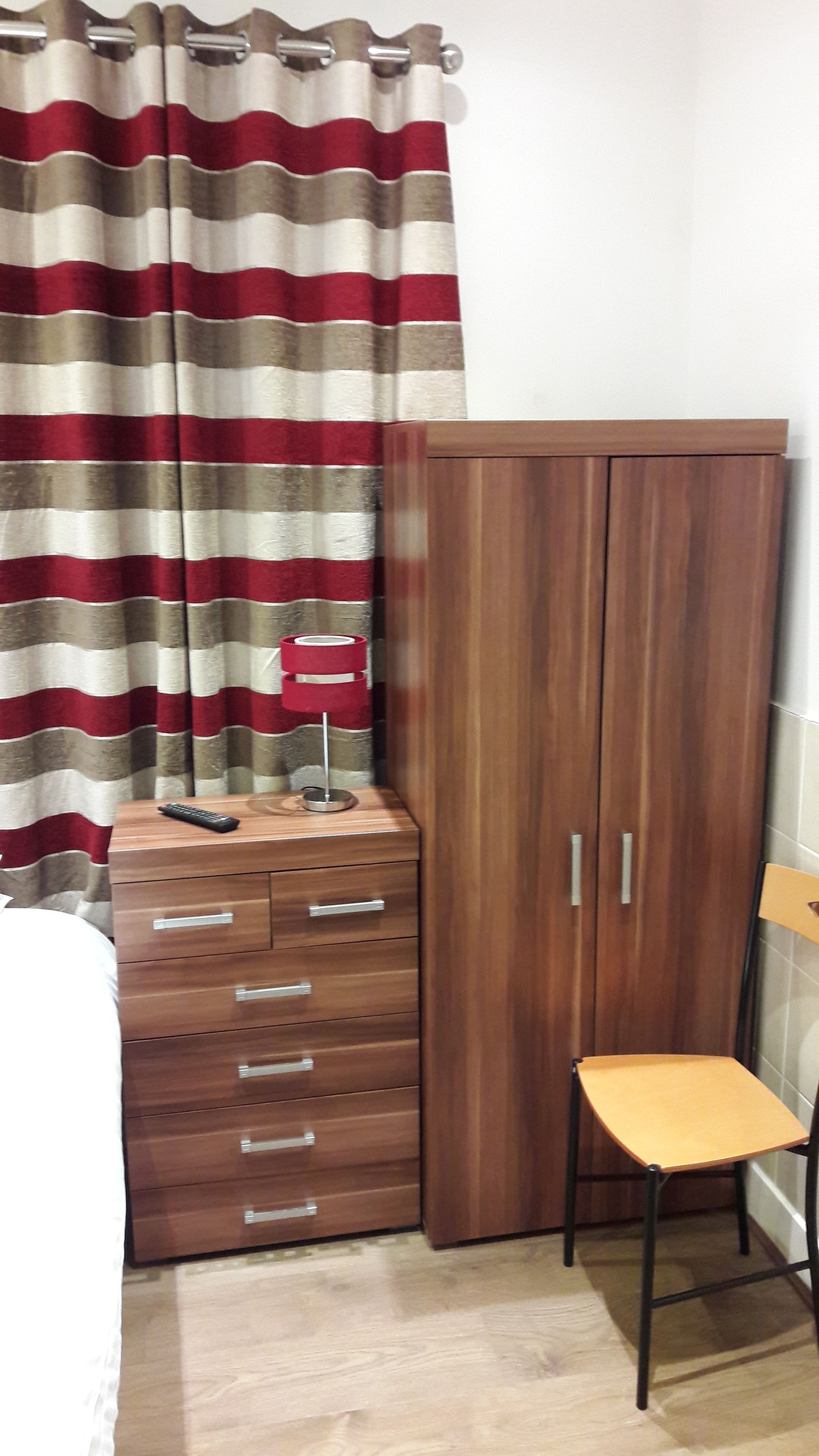 A bedroom with a wooden wardrobe, a small wooden dresser, a chair, and a bed.