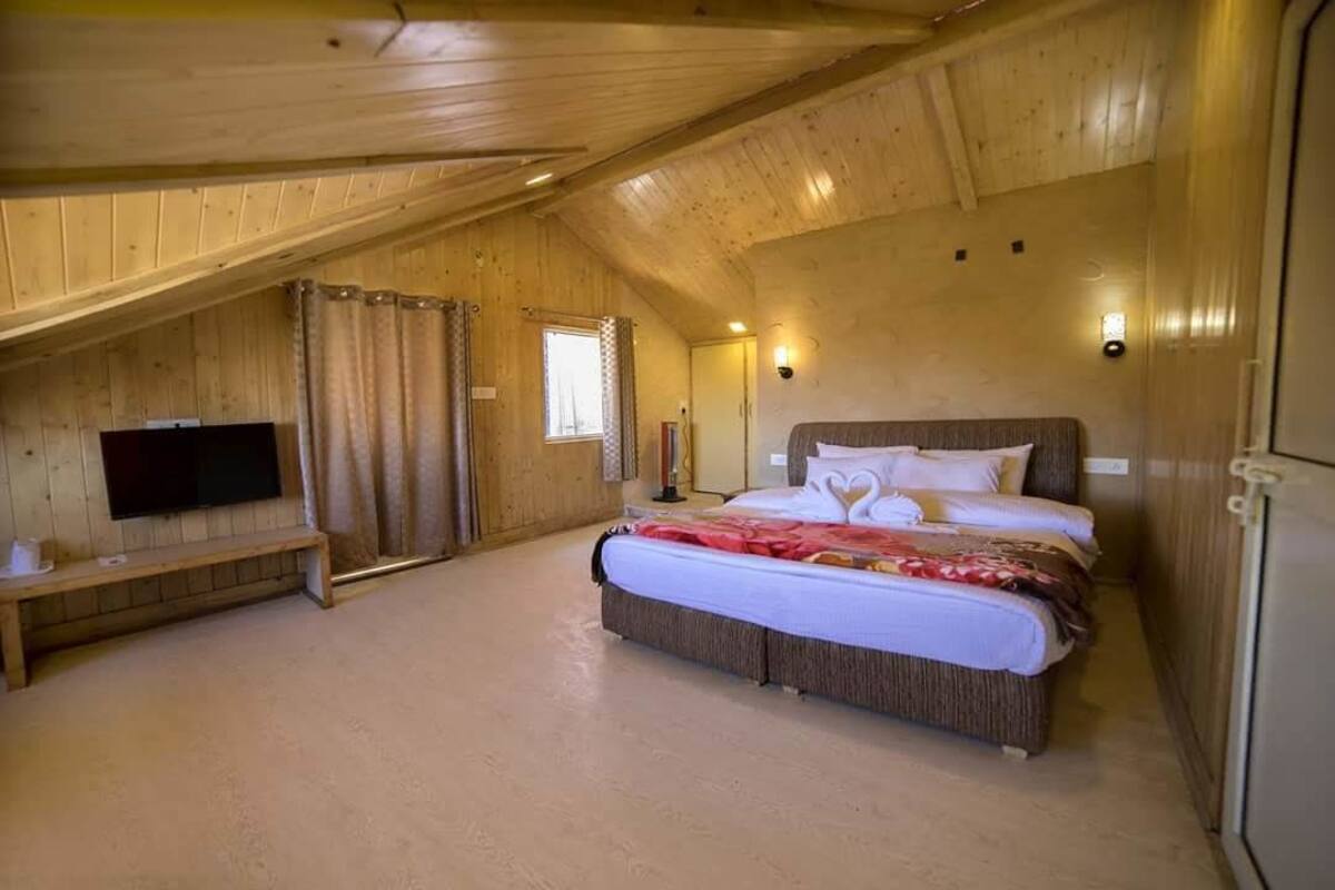 A bedroom with a wooden ceiling, a bed with a red blanket, a bench, a television, and a window with curtains.