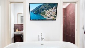 Ett badrum med ett stort badkar, en tv som är monterad ovanför och visar en kustvy, samt en kaklad duschhörna.