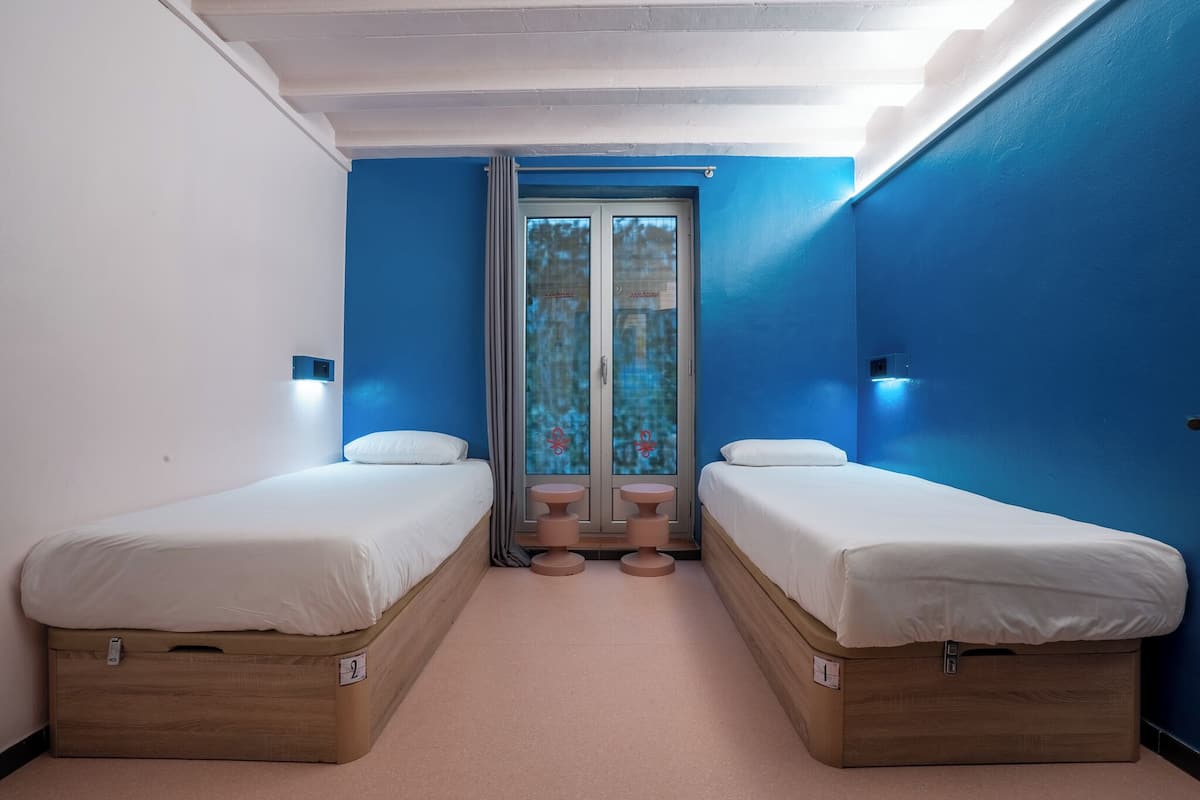 Een kamer met twee éénpersoonsbedden, een raam met gordijnen en een tafeltje tussen de bedden.