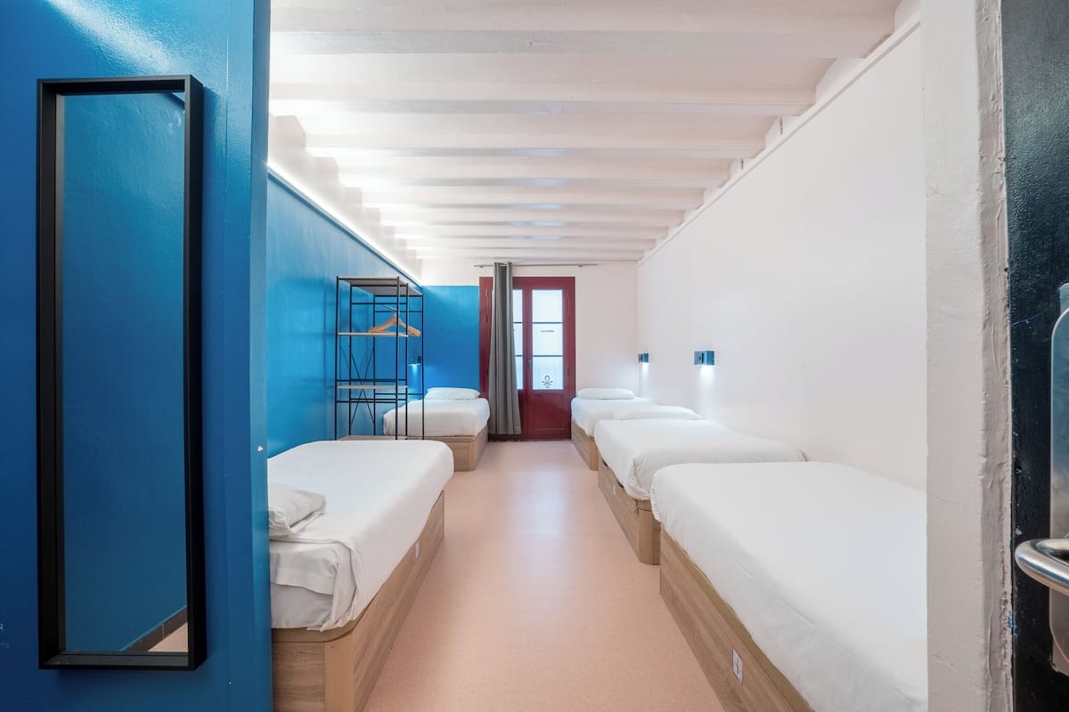 Hotelkamer met een gang die naar een deur leidt, meerdere bedden, een blauwe wand en een houten vloer.