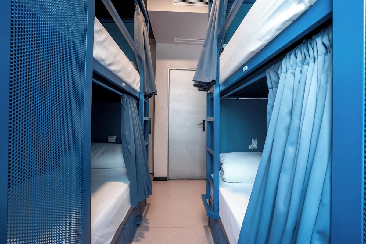 Een smalle gang met stapelbedden aan beide zijden, een deur aan het einde en een ventilatiesysteem aan het plafond.