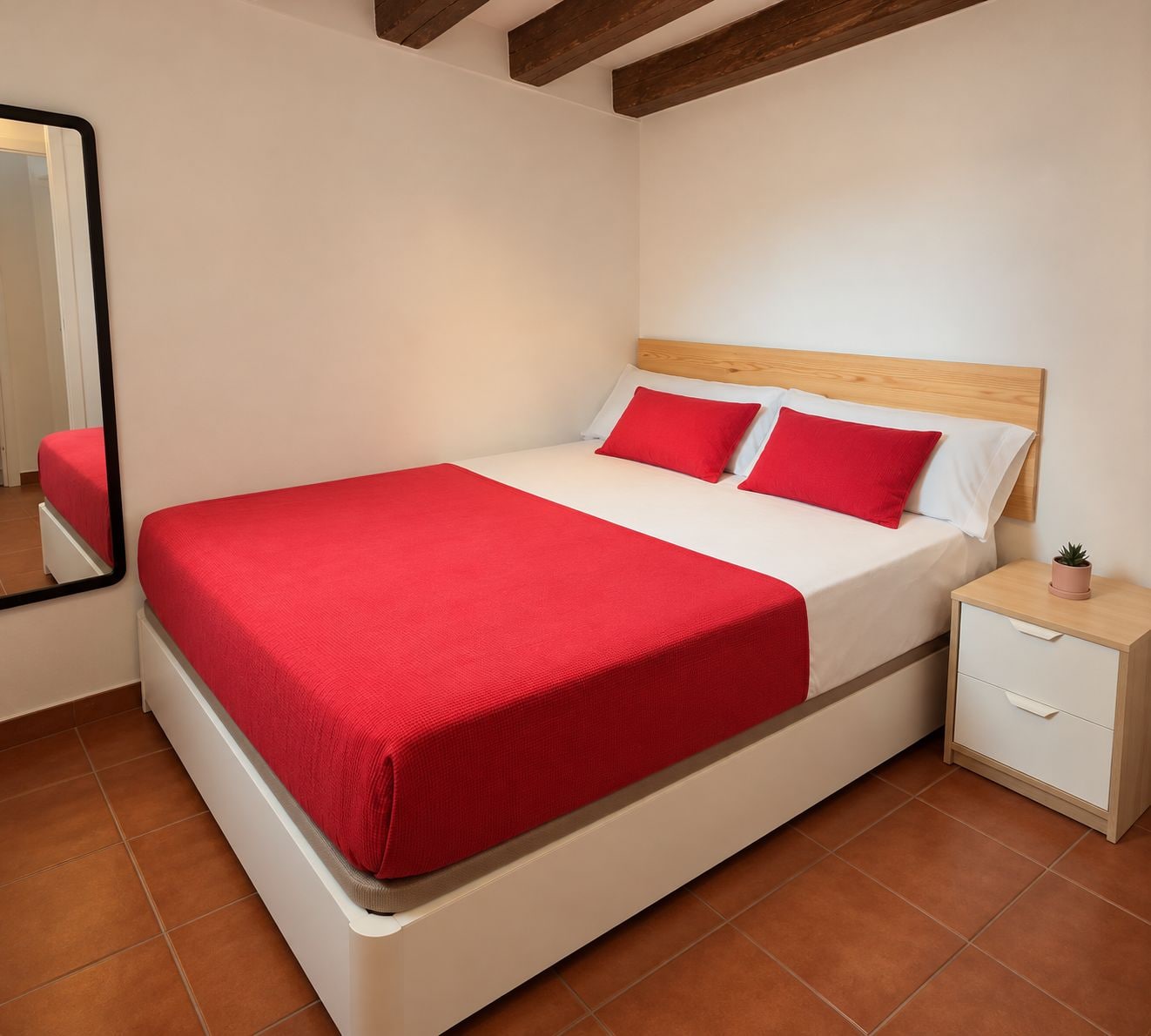 Ein Bett mit roter Bettdecke und Kissen, ein Nachttisch mit Pflanze und ein Spiegel an der Wand.
