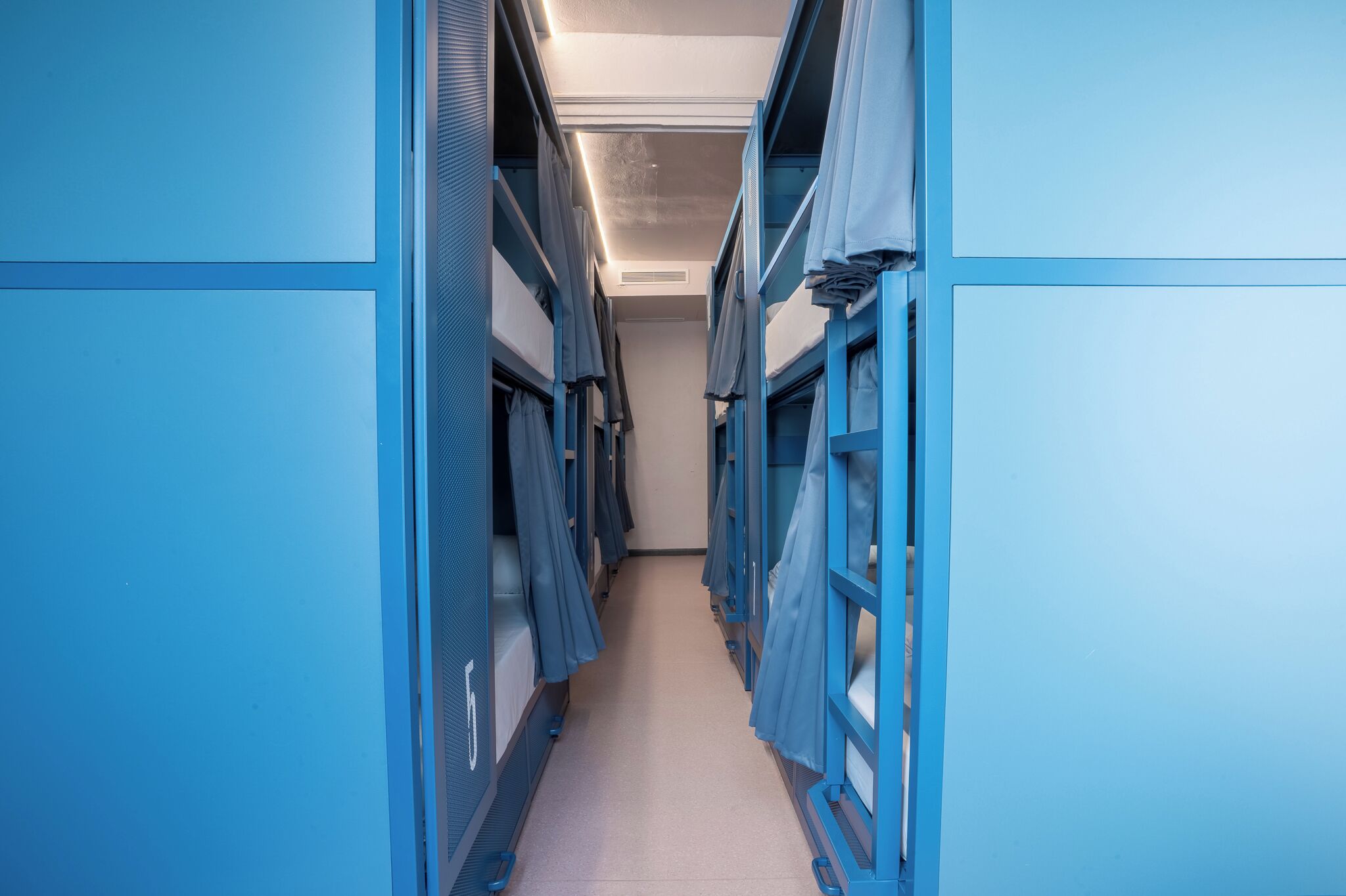A narrow corridor with bunk beds on either side, a tiled floor, and a ceiling with recessed lighting.