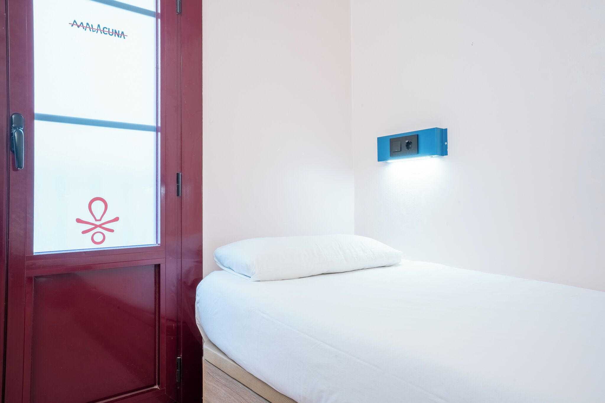 Una habitación con una cama, una puerta roja y una lámpara fijada a la pared.