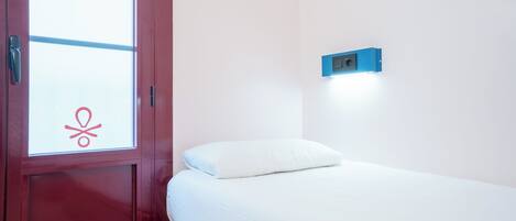 Una habitación con una cama, una puerta roja y una lámpara fijada a la pared.