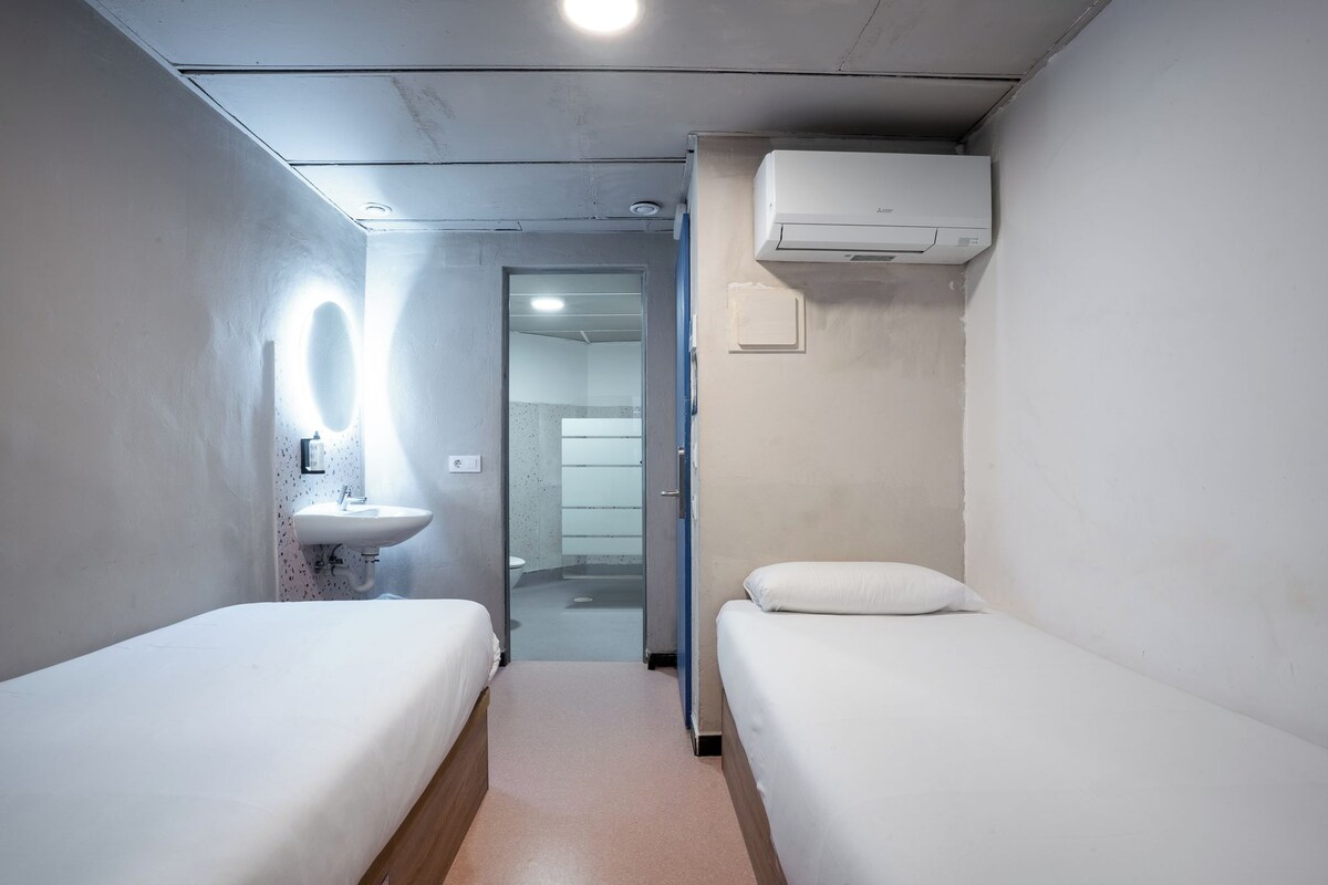 Une petite chambre d’hôtel moderne, dotée de deux lits simples, d’un lavabo intégré et d’un miroir.