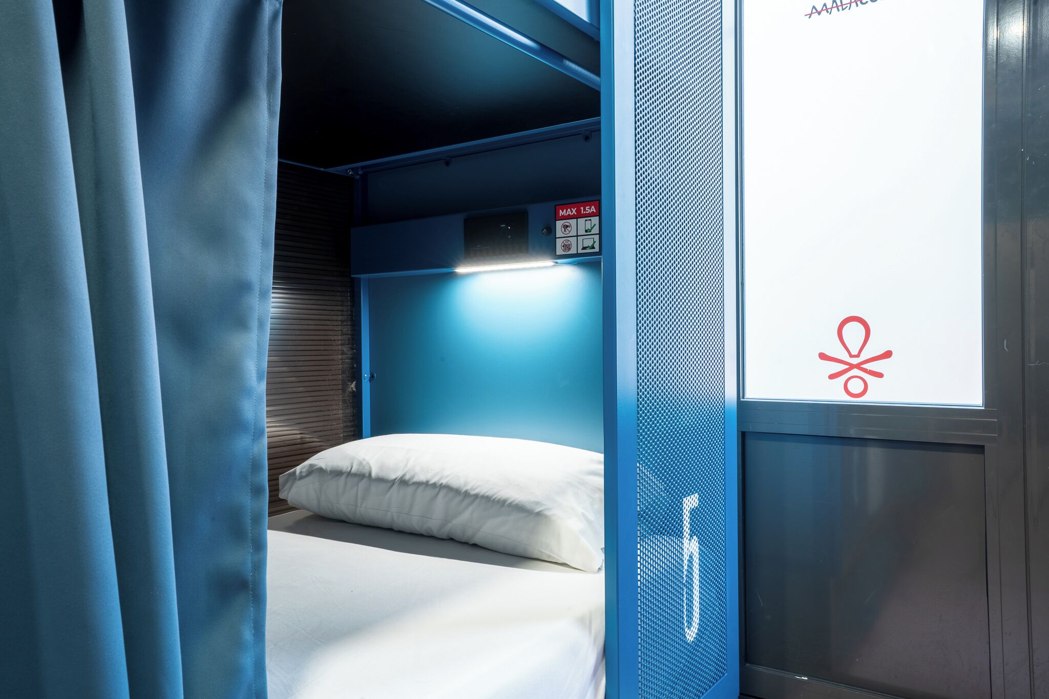 Un compartiment de train compact et bien éclairé, avec un lit simple, un mur bleu et une fenêtre ornée d’un symbole rouge.